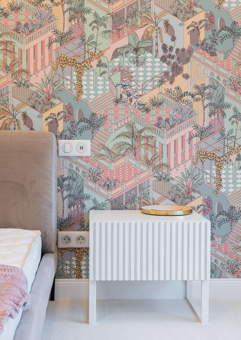 Bedroom wall covered in a pastel illustrated wallpaper of terraces and palms beside a gray upholstered bed and a white fluted nightstand
