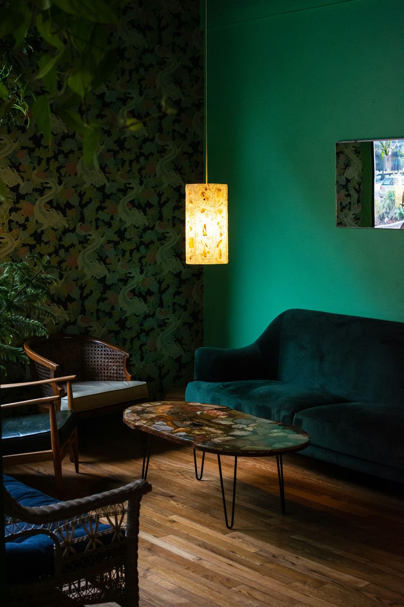 Moody sitting room with a dark botanical wallpaper, an emerald velvet sofa, cane armchairs and a glowing pendant lamp