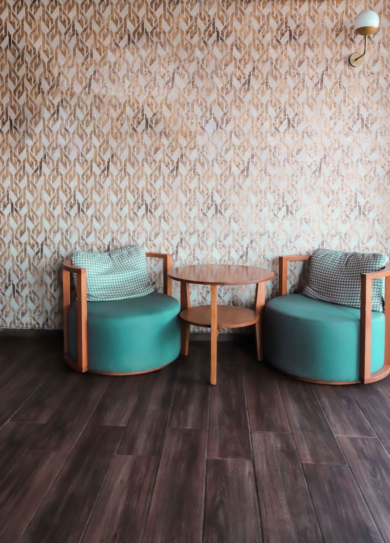 Two teal tub armchairs and a round wooden table in front of a geometric copper-and-white wallpaper on a dark plank floor