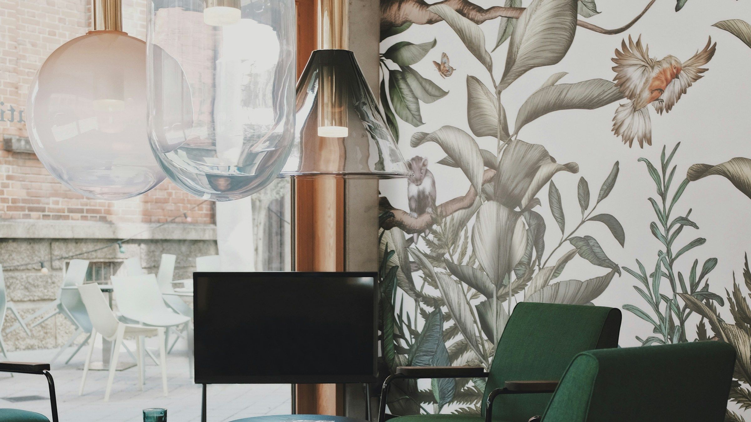 Lounge interior with a floor-to-ceiling botanical wallpaper mural, deep green armchairs, round side tables and glass pendant lights