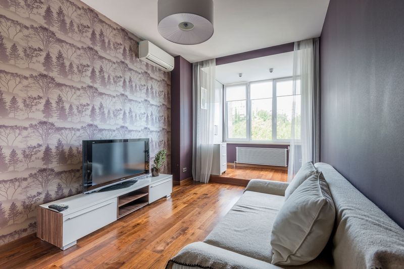 Long living room with a lilac misty-forest wallpaper wall, a white media console, a gray sofa and a bright window at the far end