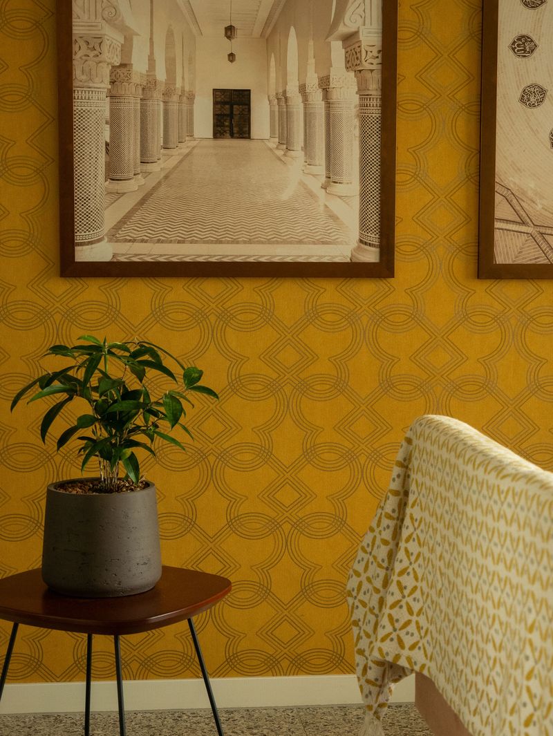 Ochre trellis-pattern wallpaper behind framed architectural prints, a potted plant on a side table and a patterned throw