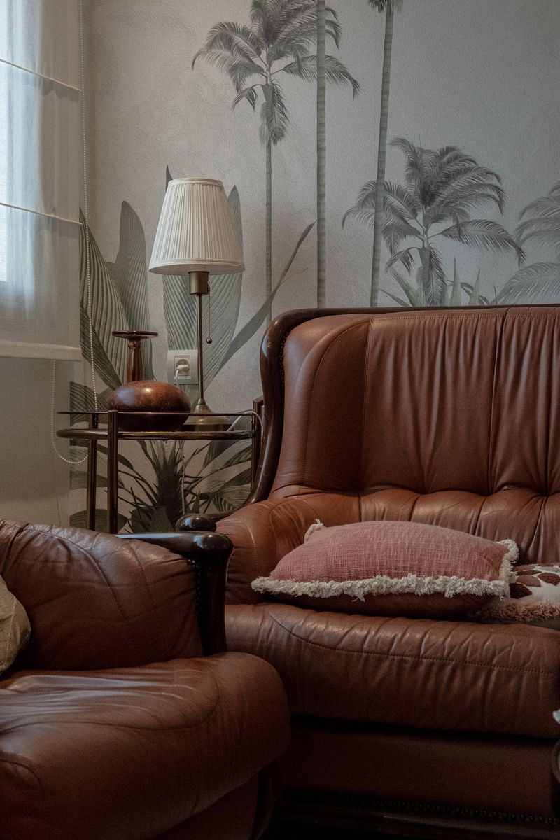Sitting area with a soft gray palm-tree wallpaper mural behind brown leather armchairs and a pleated brass floor lamp