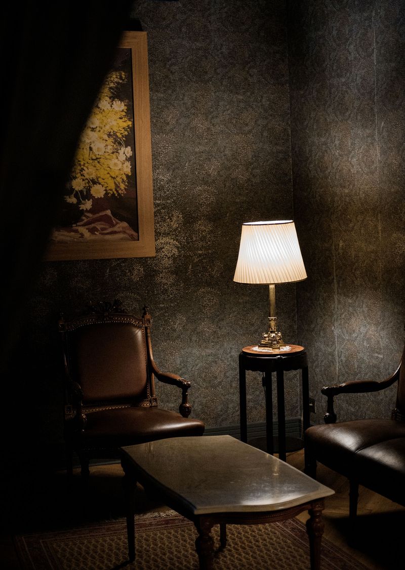 Traditional sitting room with charcoal damask wallpaper, a lit table lamp, carved leather armchairs and a framed floral painting